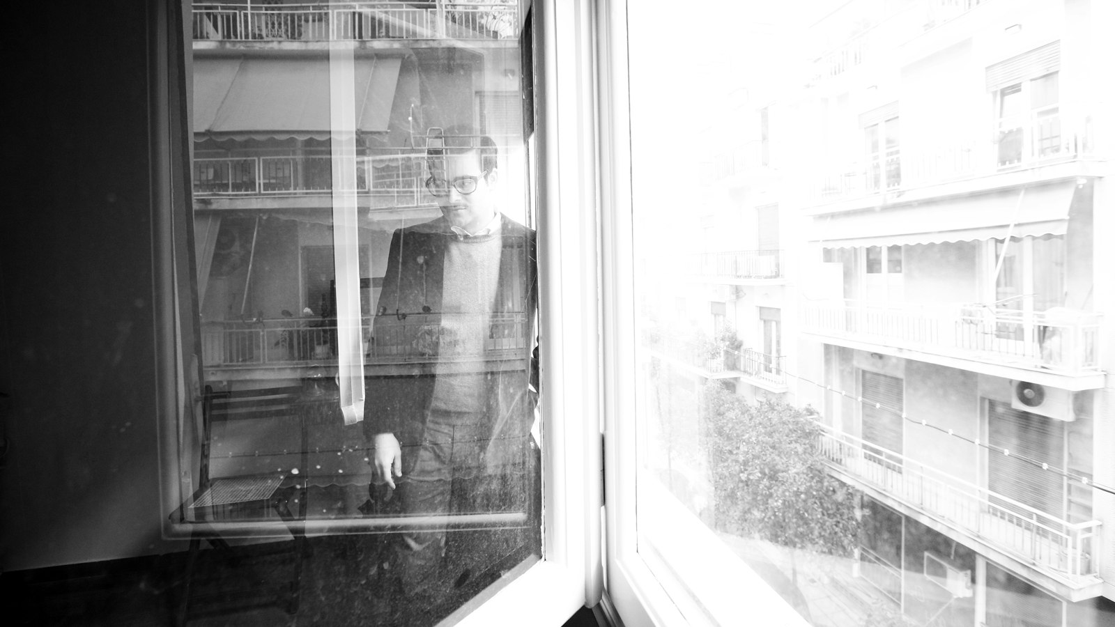 Black-and-white campaign image: the performer seen through the window of an Athens flat
