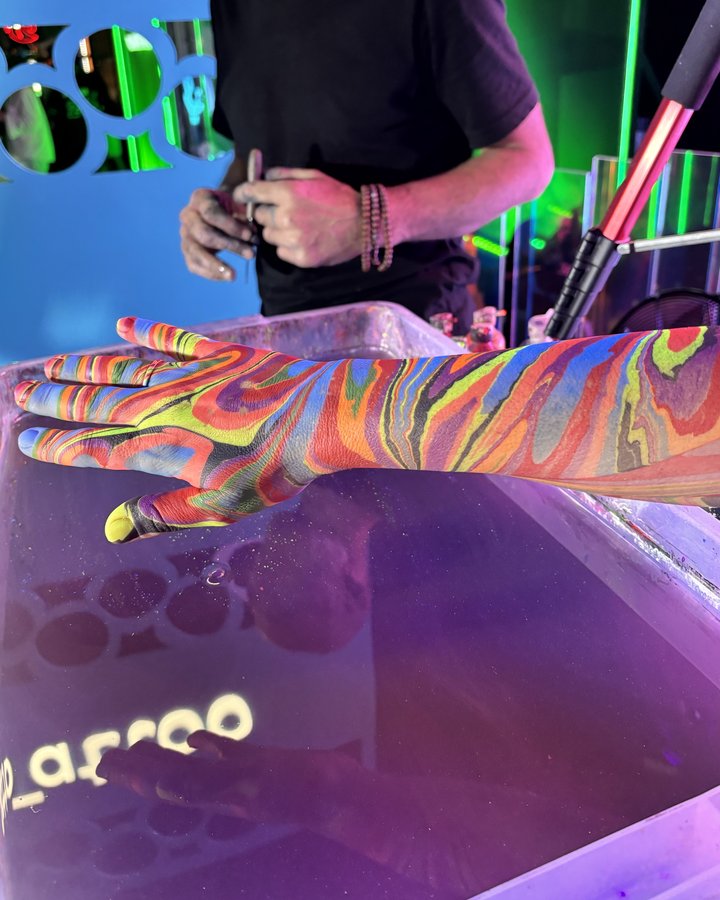 An arm and hand covered in body marbling in red, yellow, blue and green, held over the tray