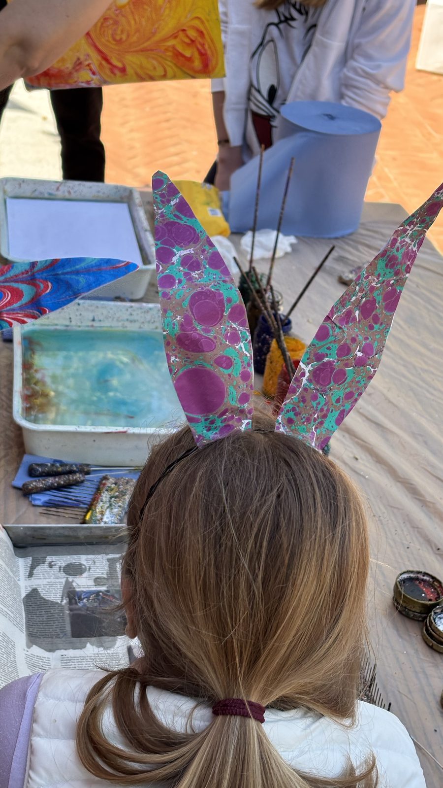 A girl in pink and green marbled bunny ears at the workshop table, beside trays of paint
